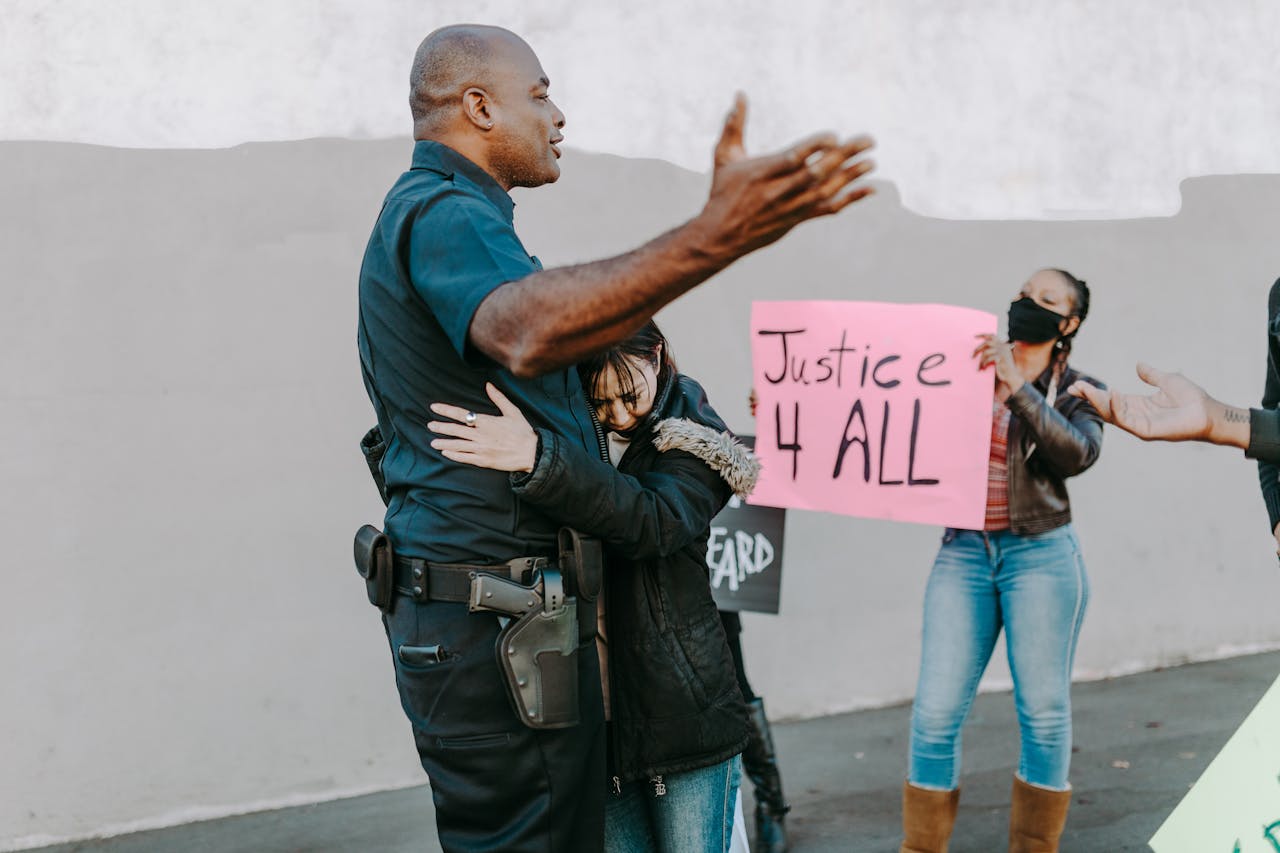 A police officer shows empathy by hugging a young girl during a peaceful protest, as a nearby woman holds a sign that reads "Justice 4 All."