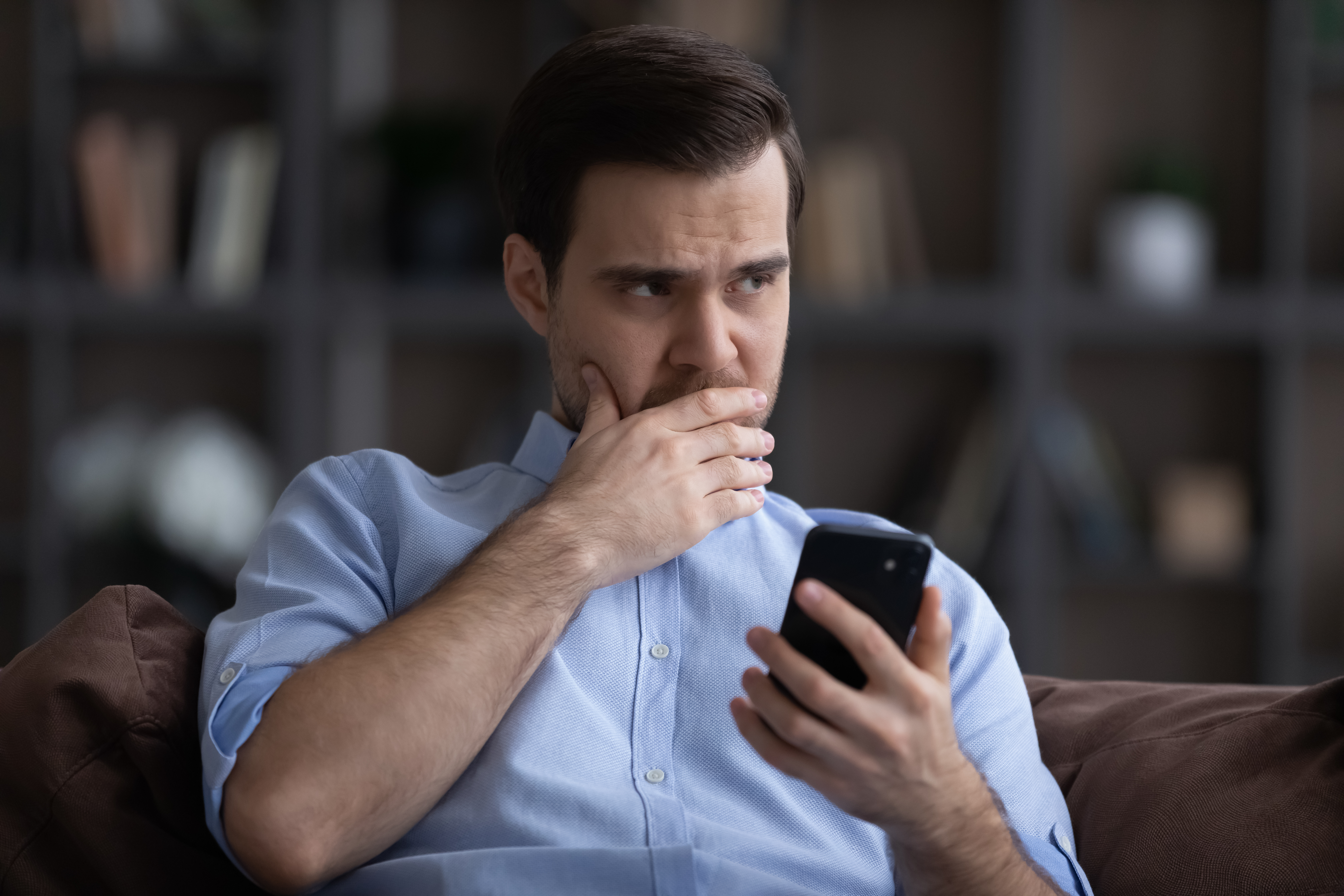 Man sitting on a couch holding a phone with a worried expression, appearing hesitant to make a decision.